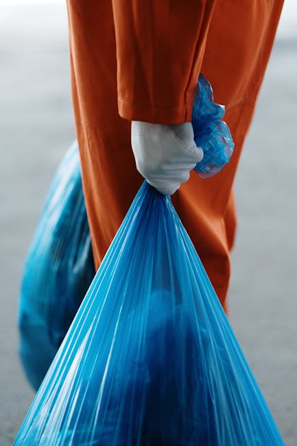 A narrow urban alleyway strewn with various types of unmanaged waste, including a large, weathered, light grey garbage bag in the foreground, positioned on a dark, uneven asphalt surface. Behind the b