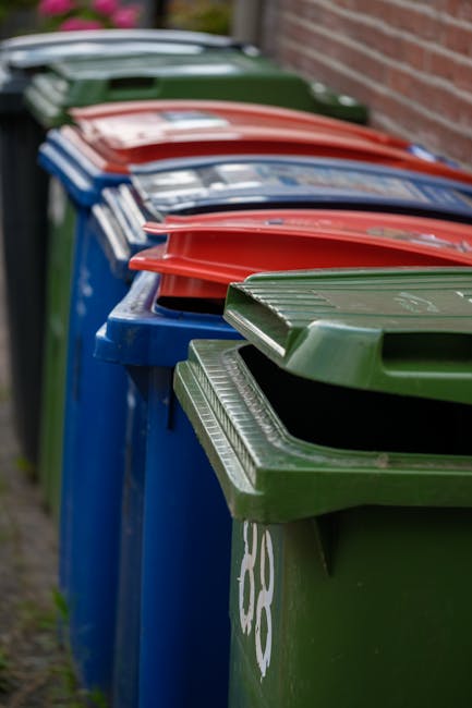 A row of large wheeled rubbish bins lined up outdoors against a brick wall, with the bins in various colours including green, blue, red, and black. The green bin at the front has a textured lid with h