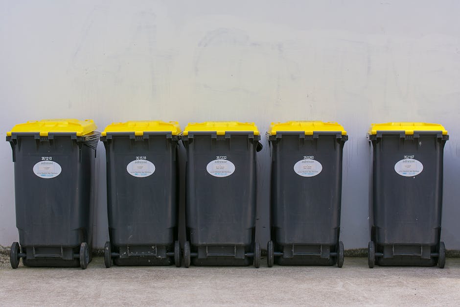 A close-up view of a cylindrical, metal cigarette disposal bin, positioned outdoors on a paved pavement surface near a building. The bin has a domed, slightly weathered lid with a matte grey finish, s